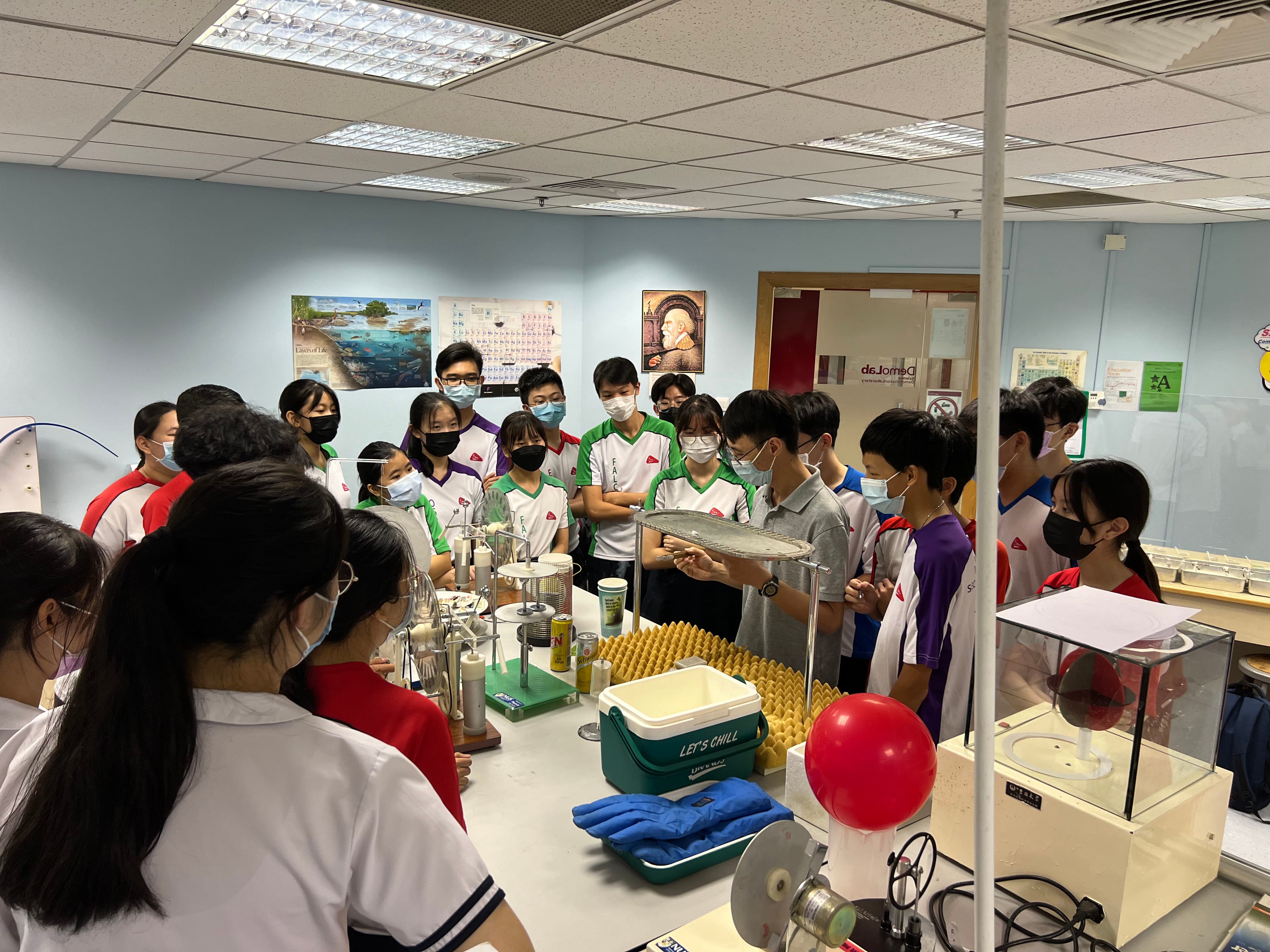 Students wearing masks gather around a science lab bench watching a demonstration with equipment and a red balloon.